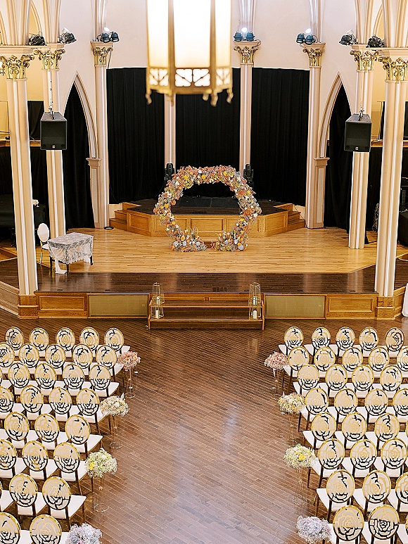 Ceremony setup for an indoor wedding ceremony with a floral arch, aisle blooms and gold lanterns, black draping in a church interior