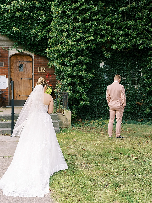 Wedding first look as the bride approaches the groom from behind, veil trailing up steps by an ivy-covered brick wall and wooden door