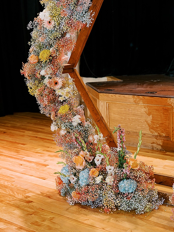 Wedding floral arch on a geometric wedding arch with baby's breath, roses, and hydrangea, set on a wood dance floor before black drapes