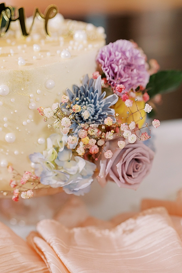 Wedding cake with floral wedding cake details, pearl sprinkles, and a gold topper on a stand beside a blush fabric drape indoors
