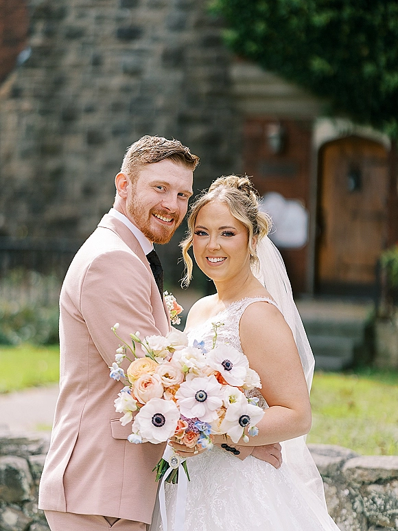 Couple portrait of bride and groom portrait with lace dress, veil, and bouquet beside a stone building with greenery and lawn