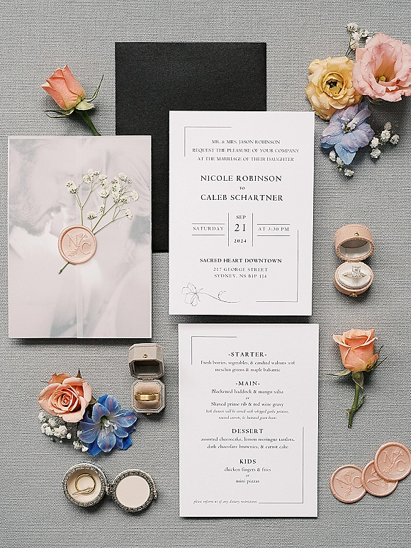 Invitation suite with modern wedding invitations on gray linen, styled with wax seals, ring boxes, bands, roses, and blue flowers
