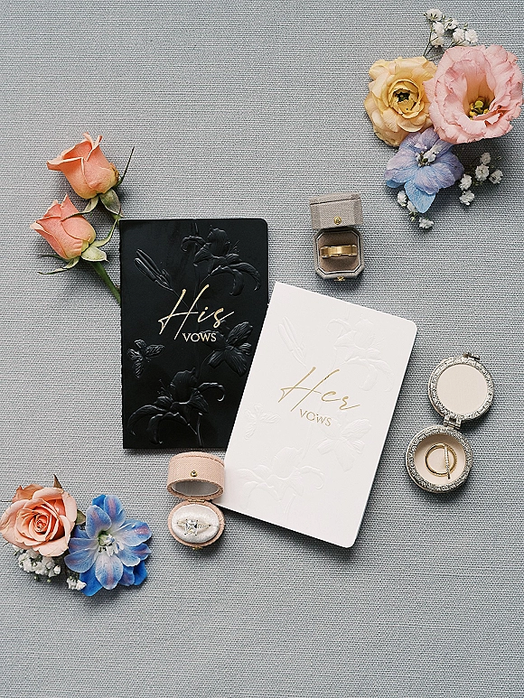 Wedding vow books in black and white with his and hers vows, ring boxes, bands and engagement ring amid pastel florals on gray fabric