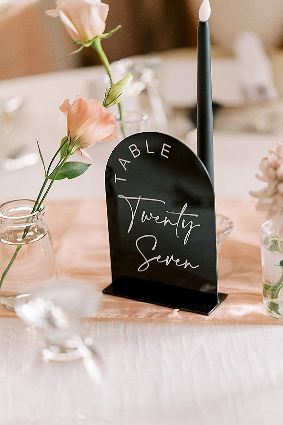Wedding table number on a black acrylic sign with calligraphy, taper candle and blush roses in glass bud vases on a reception tablescape