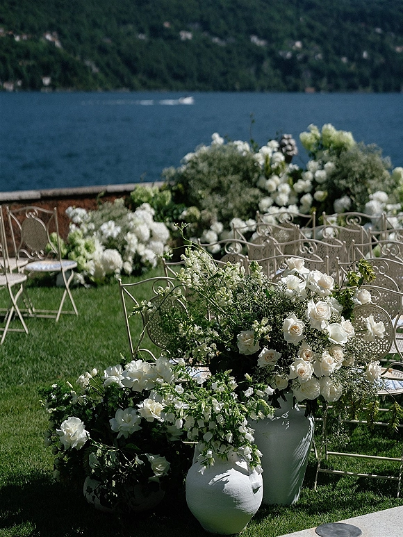 Ceremony florals with white wedding ceremony flowers in large urns and low clusters beside wrought iron chairs on a lakeside lawn