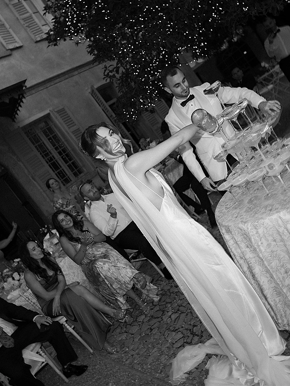 Champagne tower moment as bride and groom pour champagne into coupe glasses under string lights in an outdoor courtyard at night