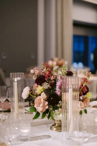 Reception tablescape with wedding table centerpiece of roses and dahlias in a compote vase, taper candles in hurricanes, and window-lit indoor room