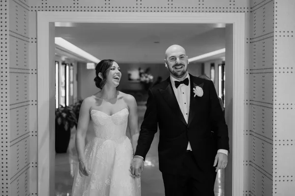 Couple portrait of bride and groom walking hand in hand, laughing in a bright hallway doorway, her strapless lace dress and his bow tie visible