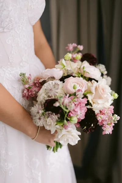 Bridal bouquet of blush and white roses with burgundy blooms and greenery held against a white lace wedding dress before dark drapery