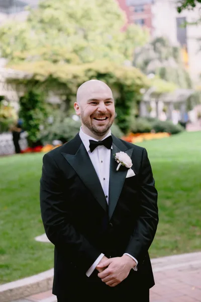 Groom portrait in a black tuxedo with bow tie and rose boutonniere, smiling on a garden lawn by an ivy-covered stone wall
