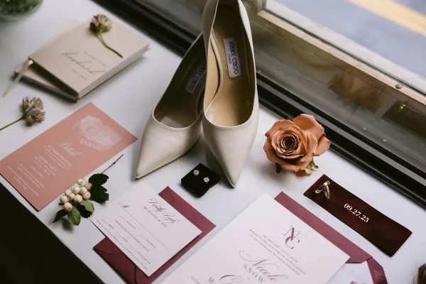 Wedding flat lay of a wedding invitation suite with bridal shoes, rings, earrings, vow book, and a rose on a window sill in soft light