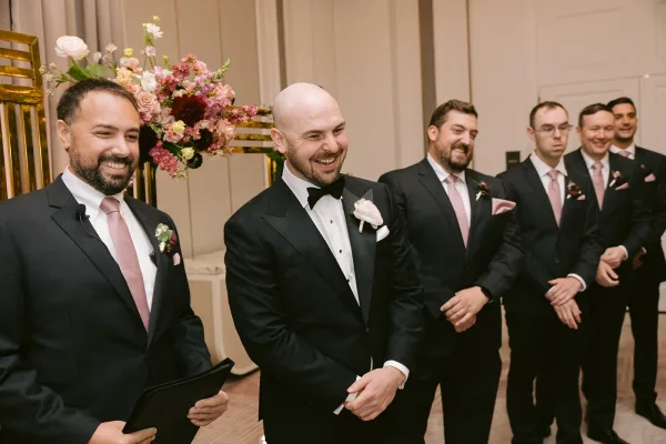 Groom portrait of a black tie groom in a black tuxedo with boutonniere, standing by paneled walls and a doorway indoors