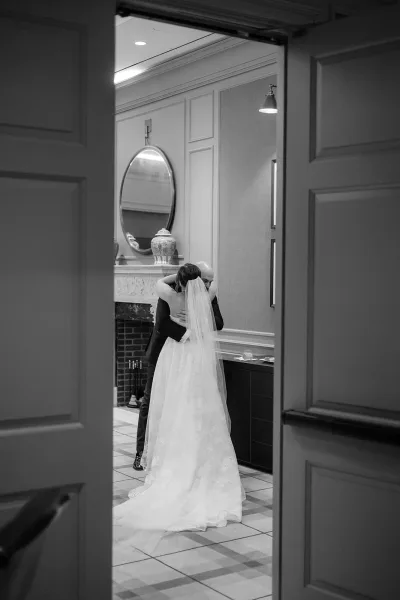 First look moment as bride in a long veil embraces the groom by a fireplace mantel in an elegant paneled hallway with mirror and sconces