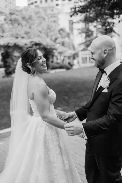 Wedding couple portrait of bride and groom laughing while holding hands, her lace gown and veil beside his tuxedo on a garden lawn