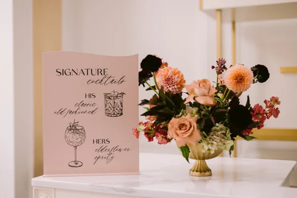 Signature cocktail sign wedding bar sign on a white countertop with a rose and dahlia arrangement in a small pedestal vase, gold-trimmed edge behind