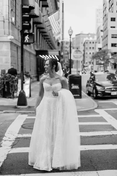 Bridal portrait in a strapless lace wedding dress, laughing while walking across a downtown crosswalk with cars and streetlights behind