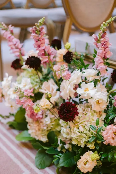 Wedding flower arrangement with pink snapdragons and white roses, hydrangeas and burgundy dahlias beside upholstered chairs on a striped rug