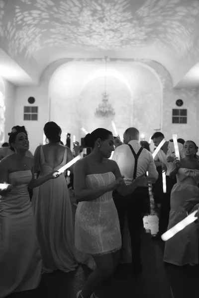 Wedding reception dancing as the bride in a strapless mini dress and wedding party wave glow sticks on a crowded dance floor under chandeliers