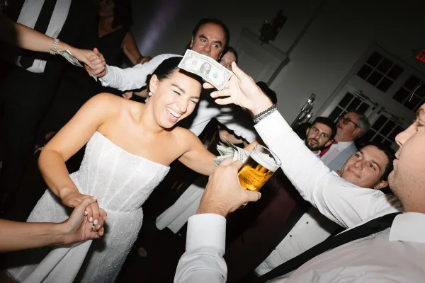 Wedding reception dancing as the bride in a strapless dress laughs while guests throw cash, with a beer glass raised in a ballroom crowd