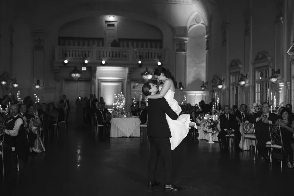 First dance lift as groom in tuxedo raises bride in strapless wedding dress under chandelier and candles in grand ballroom with guests watching