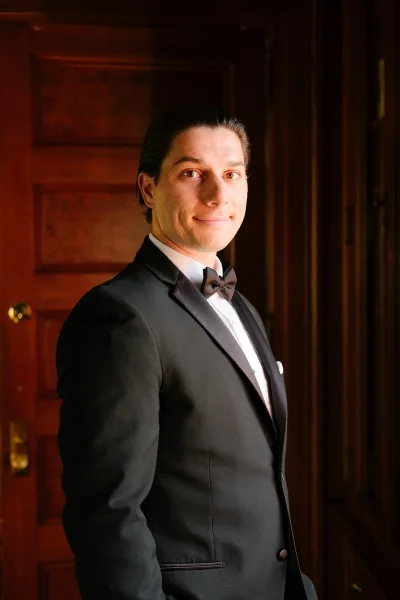 Groom portrait in a black tuxedo with bow tie and pocket square, standing by a wooden door in a warm-lit indoor hallway