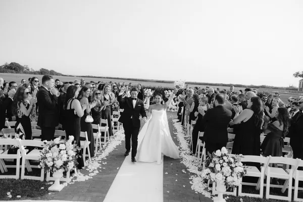 Wedding recessional as bride and groom walk the aisle, bride lifting bouquet overhead on a white runner with rose petals in an outdoor field