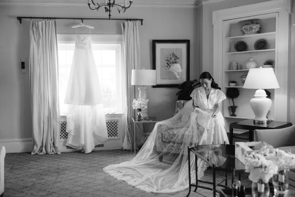 Bridal getting ready bride in a silk robe adjusts her veil by window light in a living room, wedding dress hanging nearby