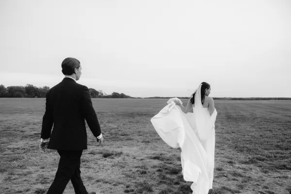 Couple portrait in a black and white wedding portrait, bride and groom walking away in an open field, her veil trailing in the wind