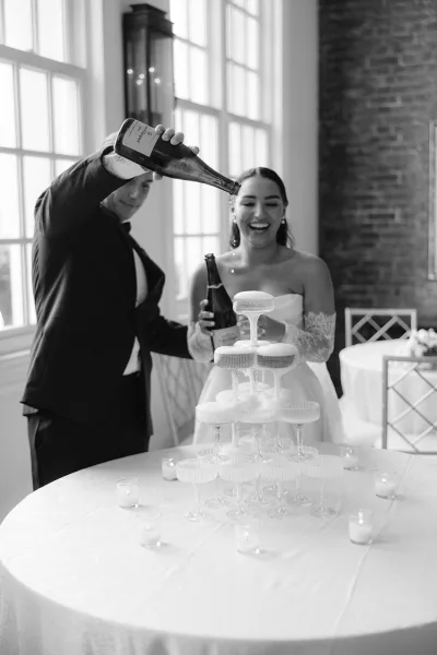 Champagne tower moment as bride in lace gloves and groom in tux pour into stacked coupe glasses by brick wall windows with votive candles