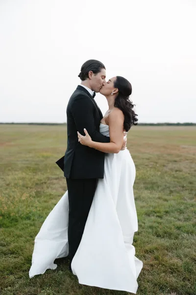 Wedding kiss portrait of bride and groom kissing in an open field under an overcast sky, her strapless dress and his black tuxedo visible