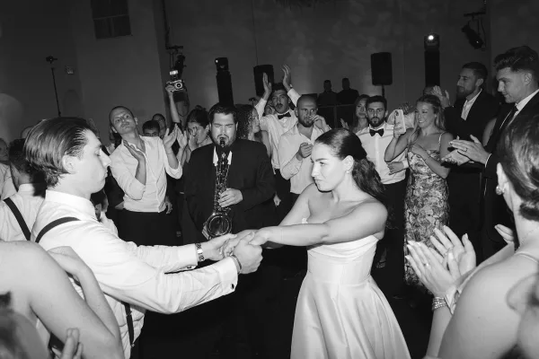 Wedding reception dancing as bride and groom in a circle dance while guests clap around them on an indoor floor under stage lights, saxophone playing