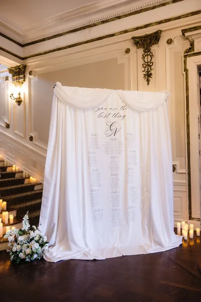 Seating chart display with a calligraphy wedding seating chart on white fabric draping, flanked by pillar candles on a grand staircase entryway