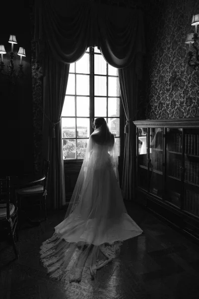 Bridal portrait in black and white of a bride in veil, lace train spread on wooden floor by a tall paned window in a library room
