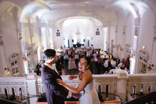 Couple portrait of bride and groom balcony pose, her strapless sparkly gown and his black tuxedo above a lit ballroom reception crowd