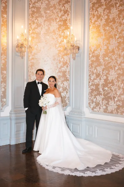 Couple portrait of bride and groom in black-tie attire, bride holding a white bouquet in front of ornate wallpaper and sconces