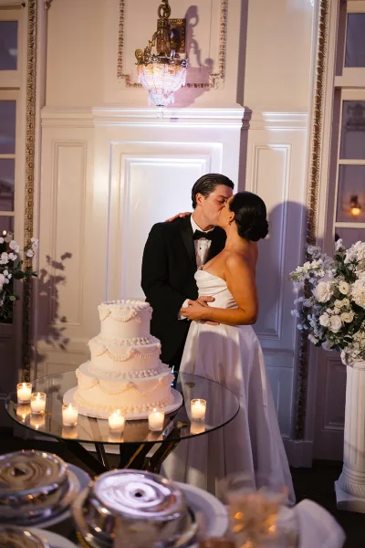 Wedding kiss portrait of bride and groom kissing beside a tiered cake on a candlelit glass table under a chandelier in an ornate room