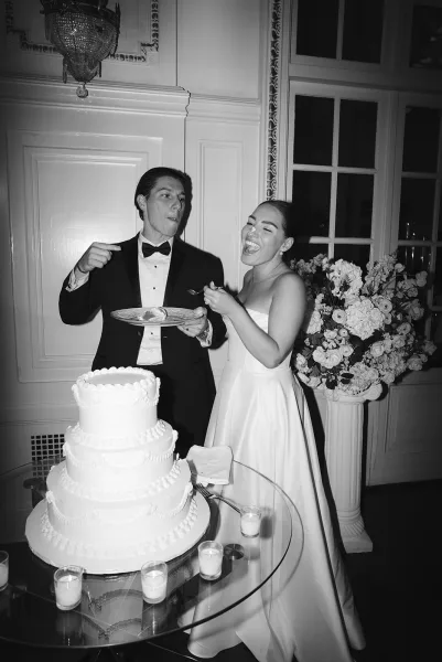 Wedding cake cutting as bride in a strapless dress and groom in a black tuxedo laugh by a three-tier cake on a glass table with candles in a chandelier-lit reception room