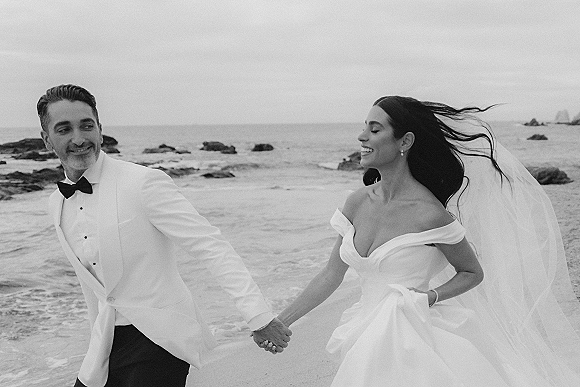 Couple portrait of a bride and groom holding hands, her veil blowing, walking along a rocky beach with waves under a cloudy sky