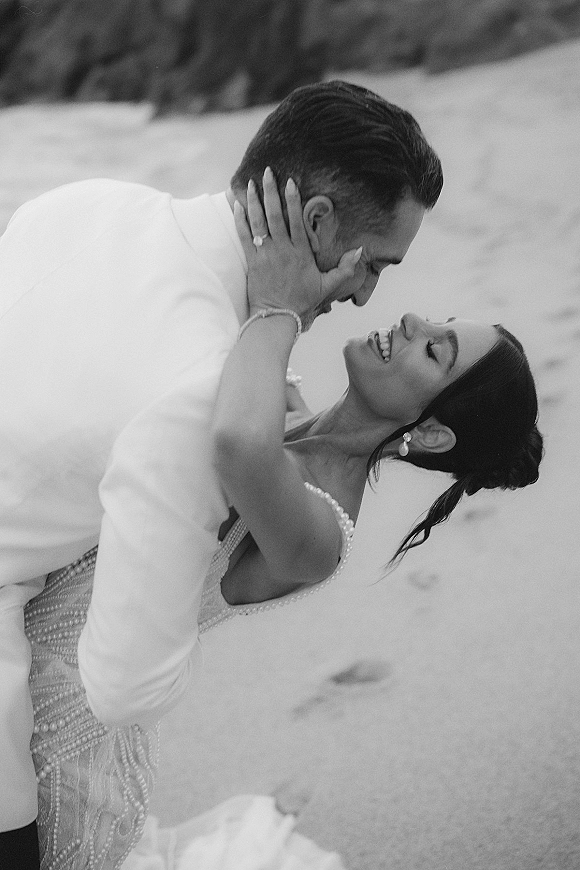 Wedding couple portrait of bride embracing groom, showing engagement ring and pearl earrings, with ocean shoreline and distant hills behind them