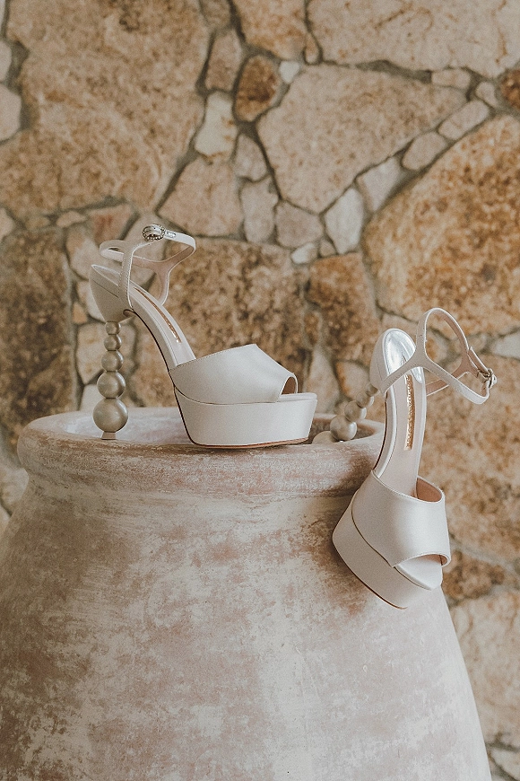 Bridal shoes, white platform heels with ankle straps and decorative heel detail set against a stone wall with a ceramic urn backdrop
