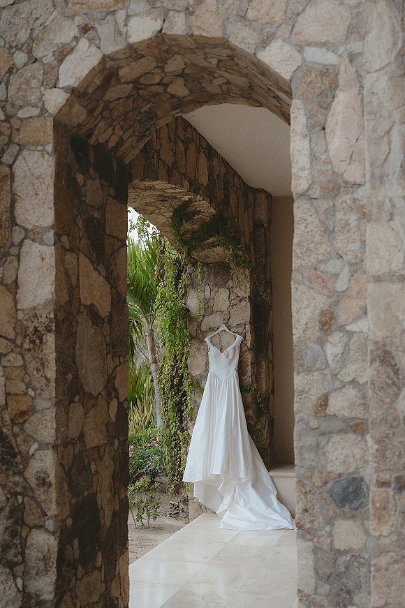 Wedding dress on hanger with spaghetti straps and button-back chapel train framed by an ivy-covered stone archway in a garden villa setting