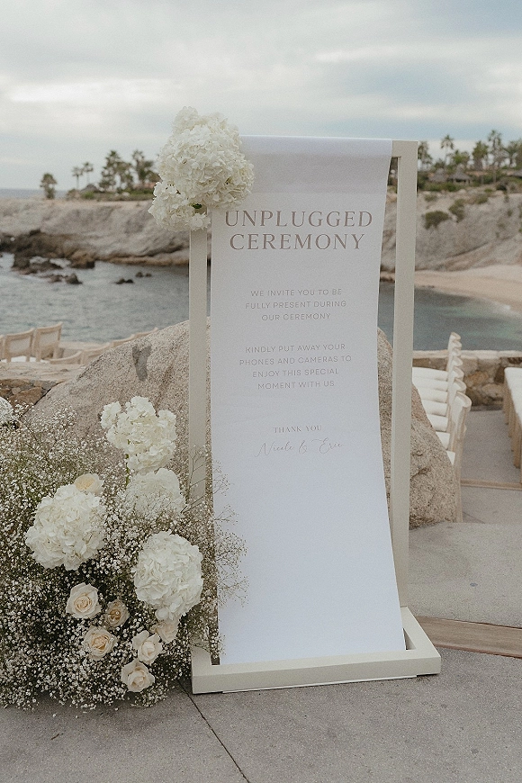 Unplugged ceremony sign with white hydrangeas, roses, and baby's breath on a stand beside a rocky ocean cove and chairs under clouds