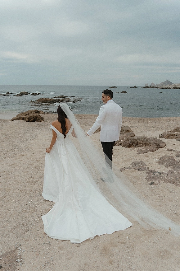 Couple portrait of bride and groom at a beach wedding couple, walking hand in hand on sand with a long veil trailing by the ocean