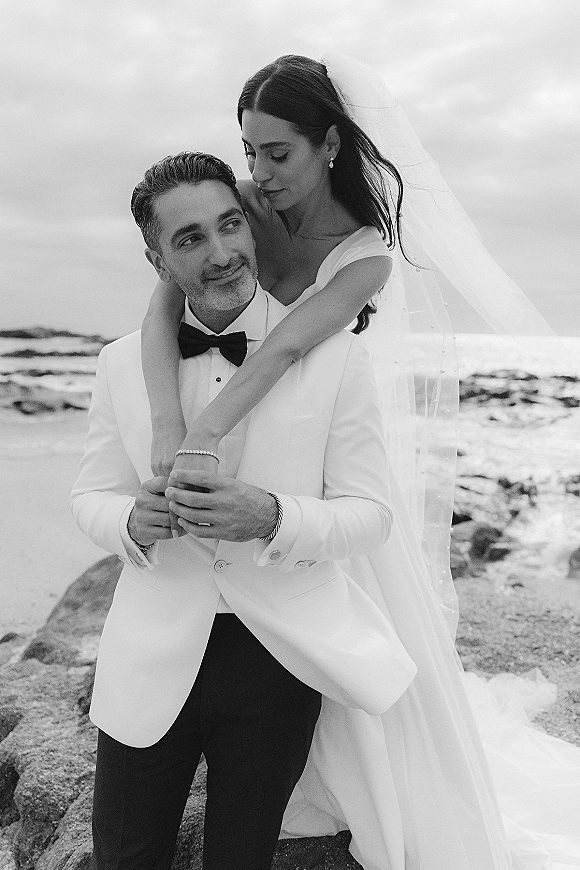 Couple portrait in a black and white wedding portrait style, bride hugging groom in white tuxedo on rocky shore with ocean behind