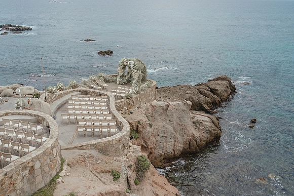 Ceremony setup for a cliffside wedding ceremony with wooden chairs and a white floral arch overlooking ocean waves and rocky coastline