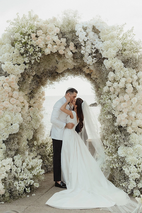 Wedding kiss portrait of bride and groom kissing beneath a white rose floral arch, her veil flowing, with ocean and sky behind