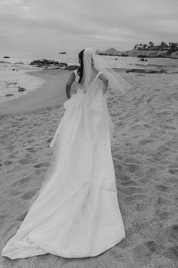 Bridal portrait of a bride on beach from behind, cathedral veil and dress train flowing over sand with ocean and rocky shoreline under clouds