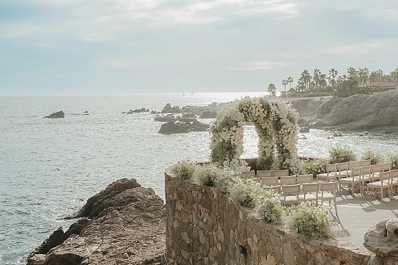 Ceremony setup for a cliffside wedding ceremony with an oceanfront white floral arch and baby’s breath aisle blooms on a stone terrace by the sea