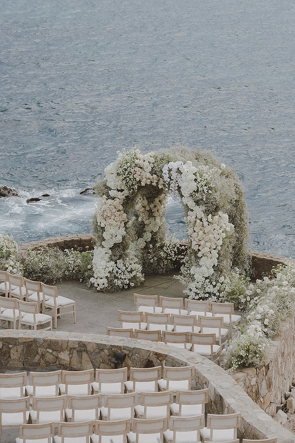 Ceremony setup for an outdoor wedding ceremony with a white floral arch and baby's breath on a stone terrace overlooking the ocean