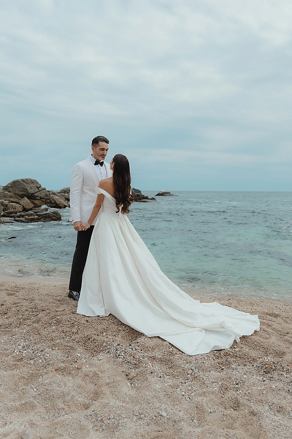 Couple portrait at a beach wedding, bride and groom holding hands on a rocky shoreline with ocean backdrop under cloudy sky